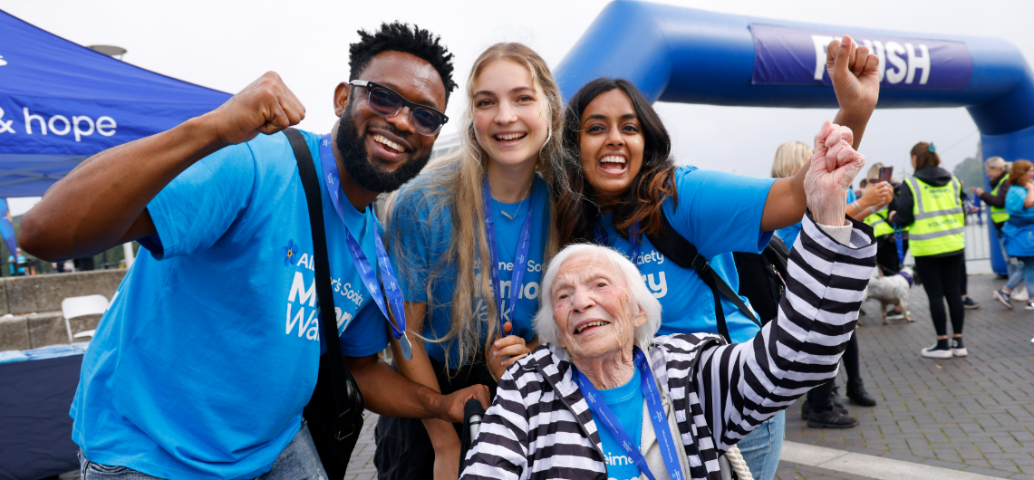 Three people stand around an older woman in a wheelchair, they all cheer.