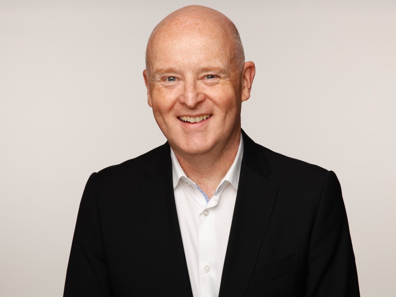 A smiling bald man against a white background wearing a white shirt and black blazer