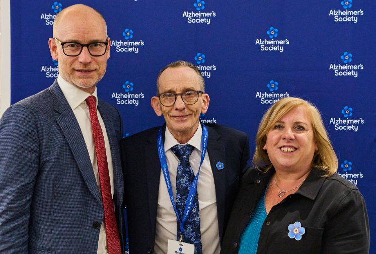 A woman and two men stand together in front of an Alzheimer's Society screen.