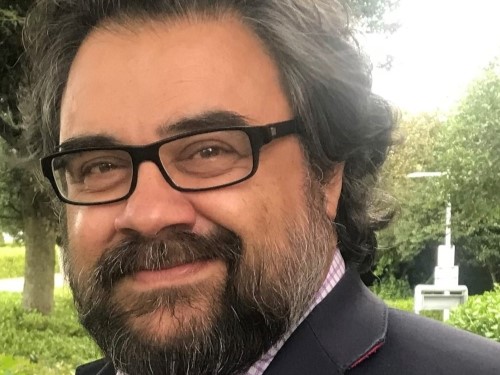 A man with black hair and a black beard, wearing glasses and smiling