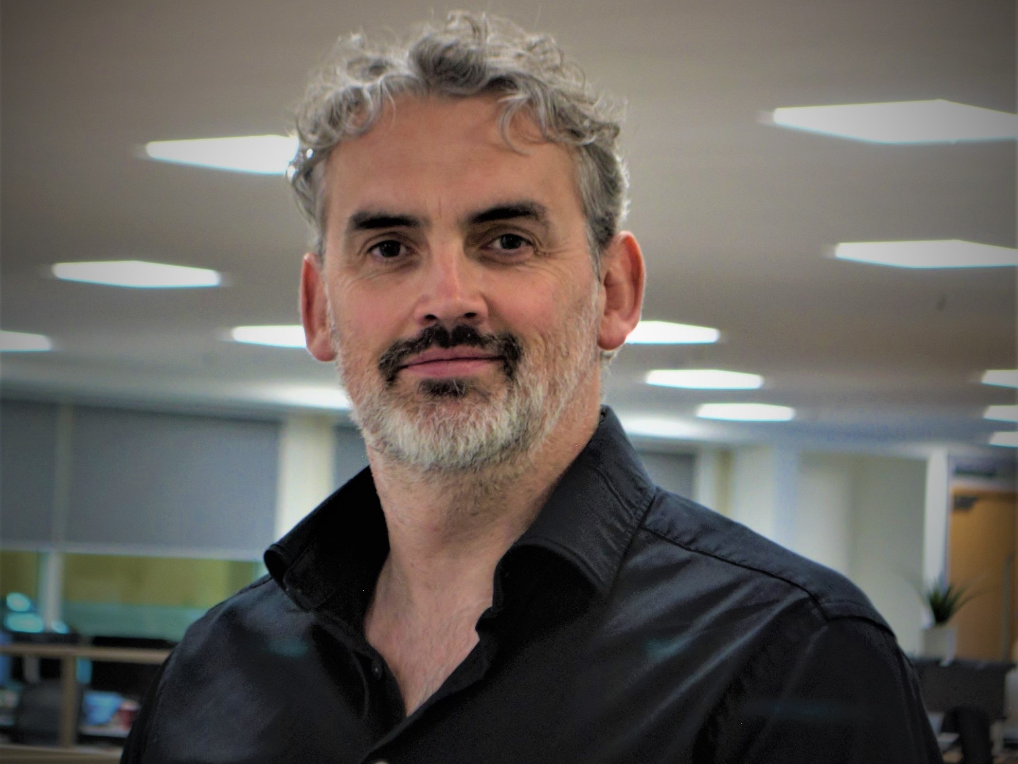 A man with grey hair and grey beard, black moustache and eyebrows, wearing a black shirt in an office