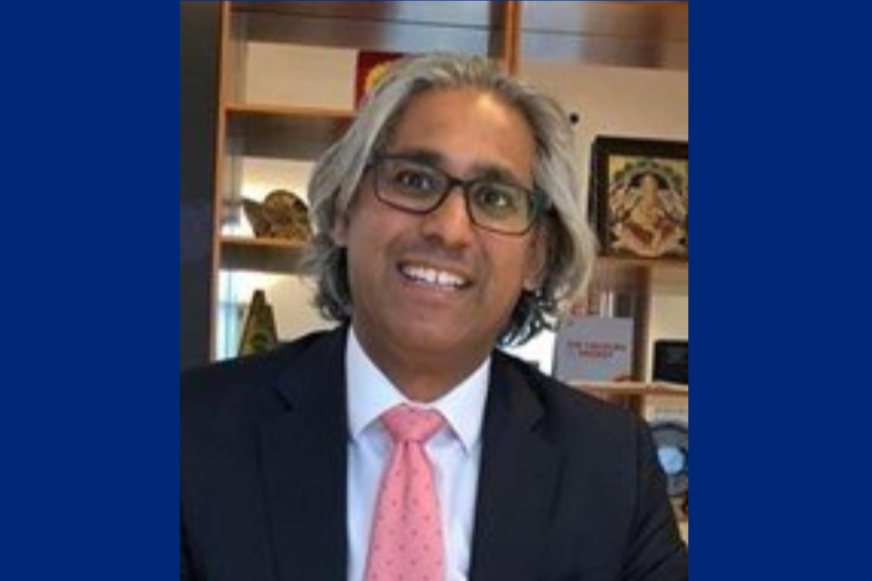 A man with grey hair, glasses, white shirt, black jacket and pink tie