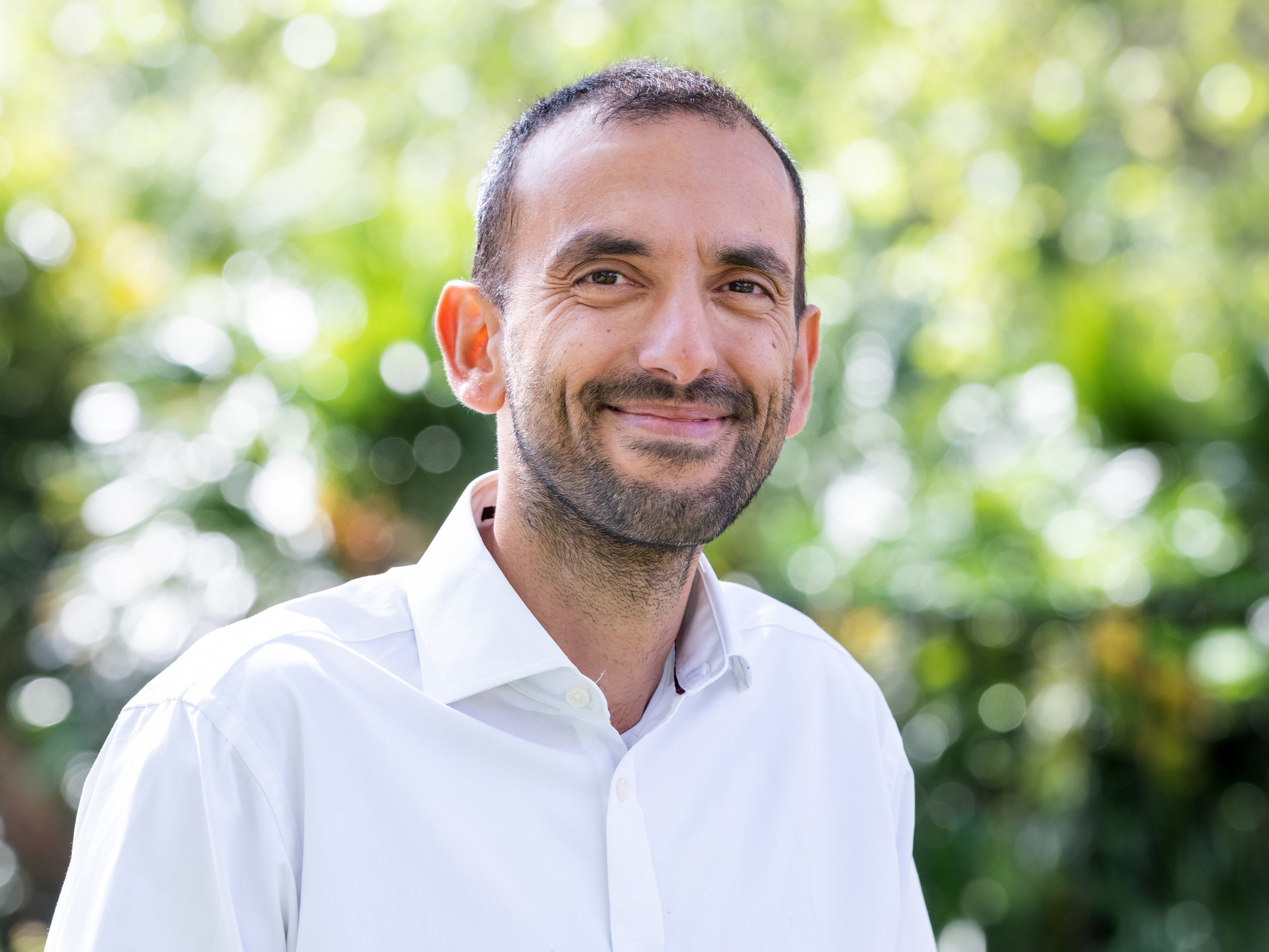 A man with short brown hair and stubble wearing a white shirt and smiling