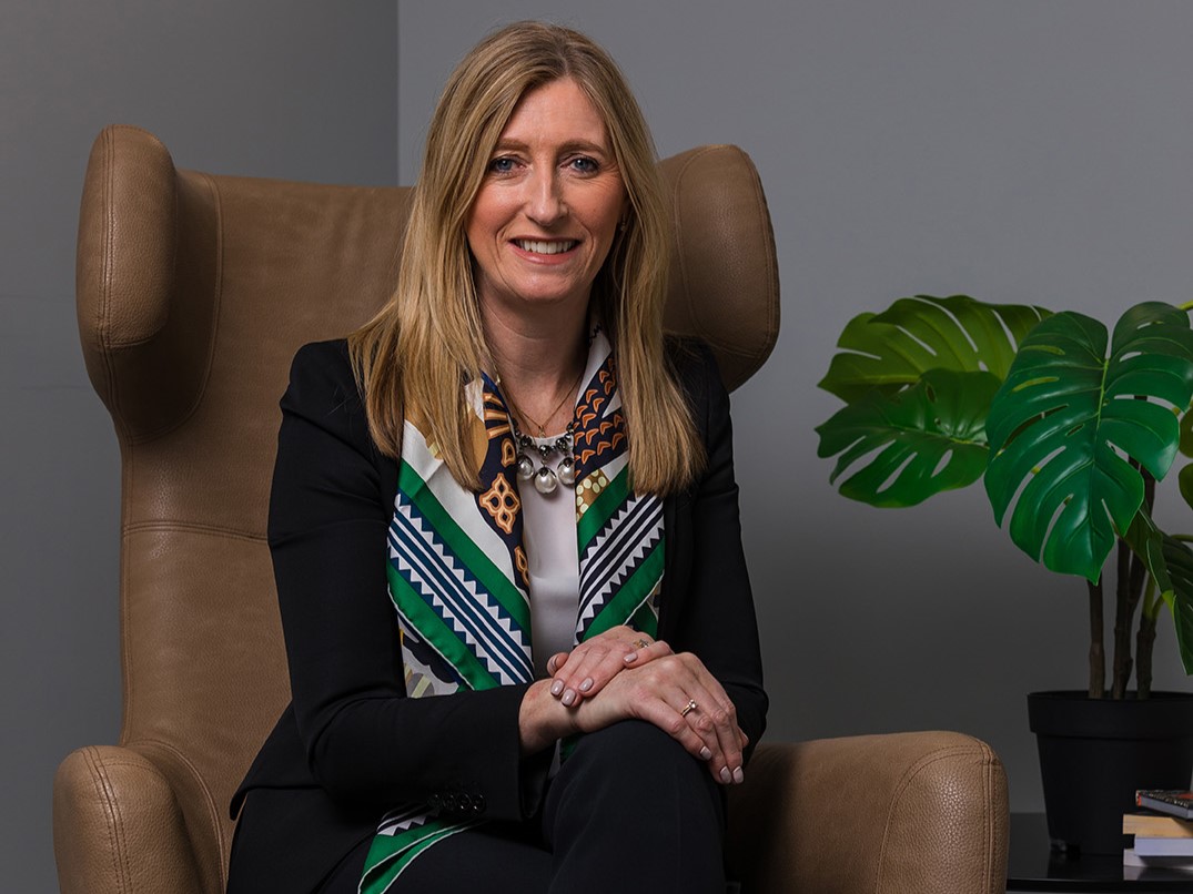 A woman with mid length blonde hair, wearing a black suit and a printed scarf, sitting on a brown chair
