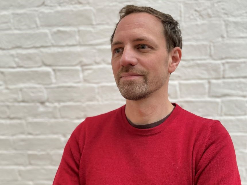 A man with short brown hair stubble, wearing a red jumper