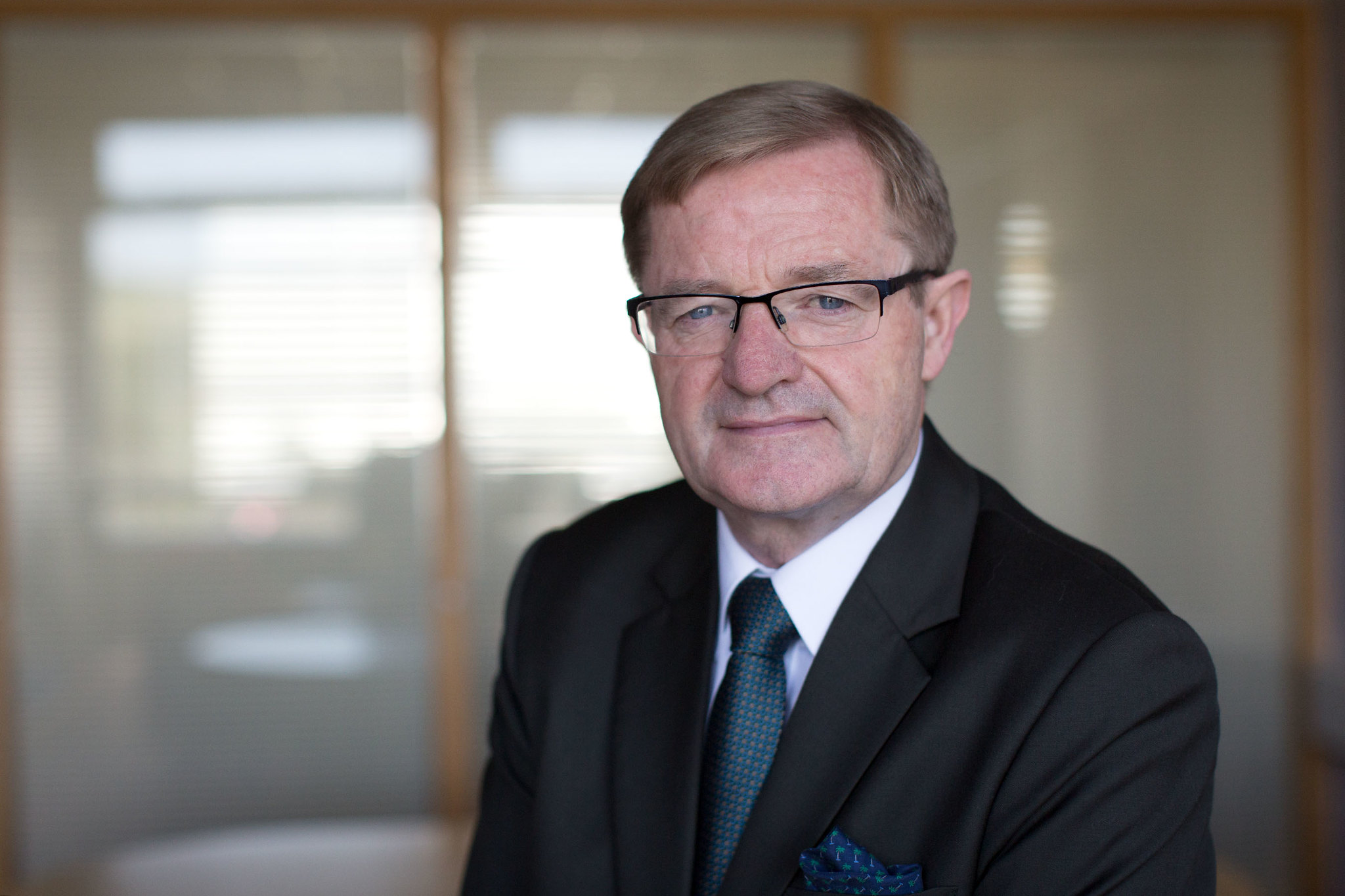 A man with short brown hair, wearing glasses and a dark grey suit with a blue tie