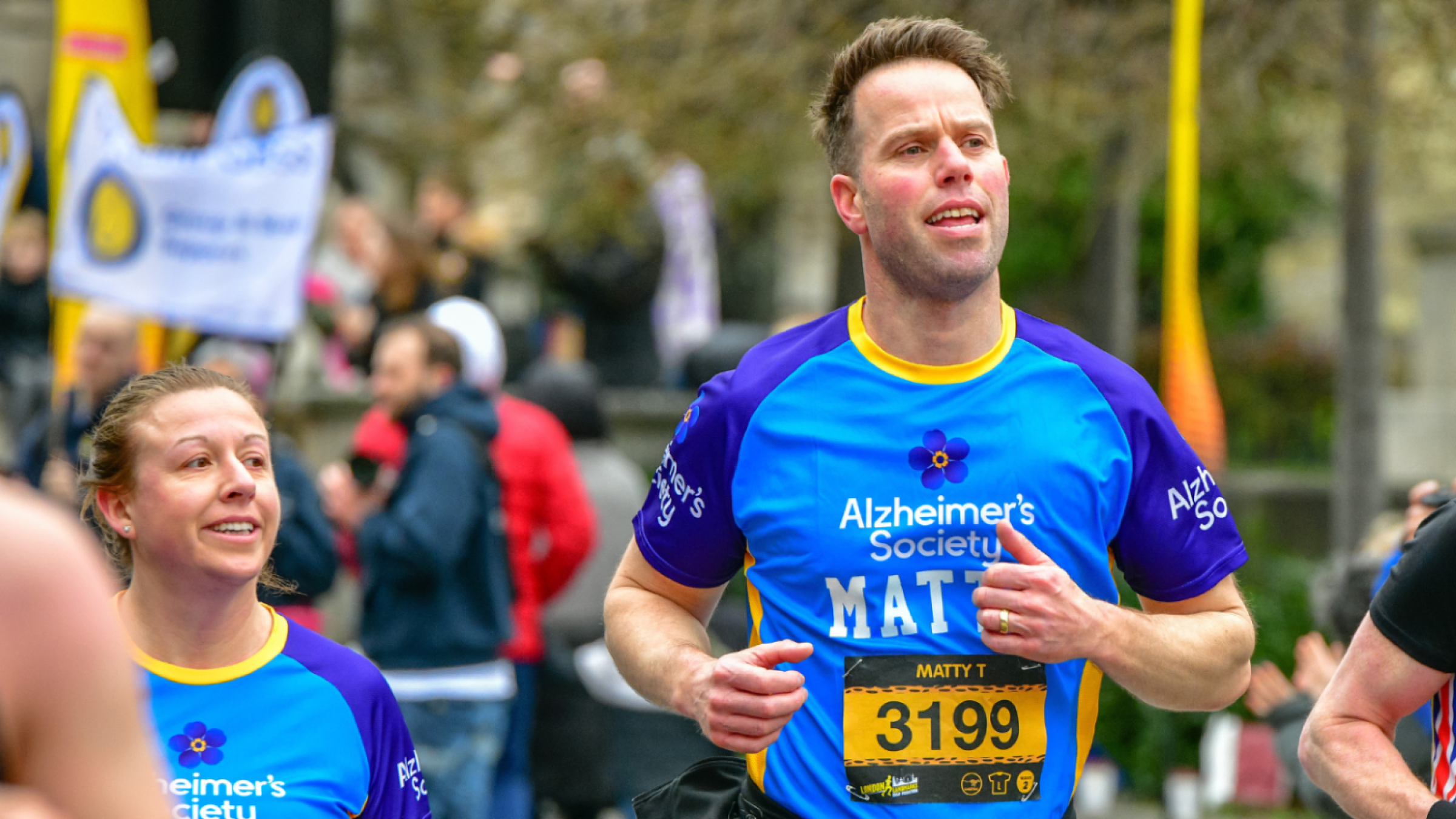 Two Alzheimer's Society runners smiling and running as they pass a crowd