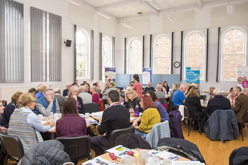 People seated at large round tables busy in conversation, the room is large and bright with high ceilings