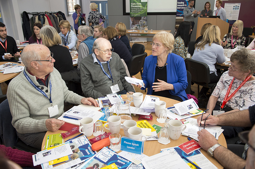 A blonde female professor sits at a round table with a group of people affected by dementia from York. She is explaining her research and they are listening