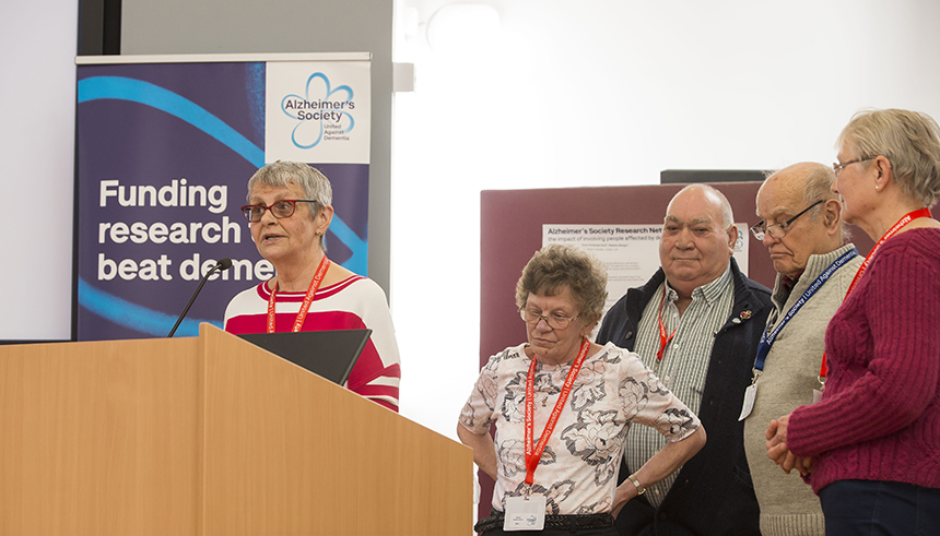 Wendy Mitchell stands speaking into a microphone at a podium while the members of the York Minds and Voices group stand behind her