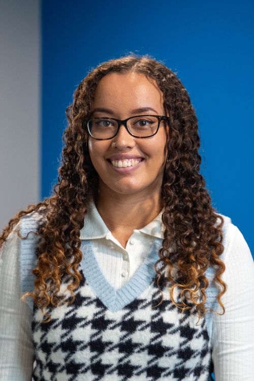 Jade, wearing a white shirt and black and white patterned jumper, with glasses, smiling