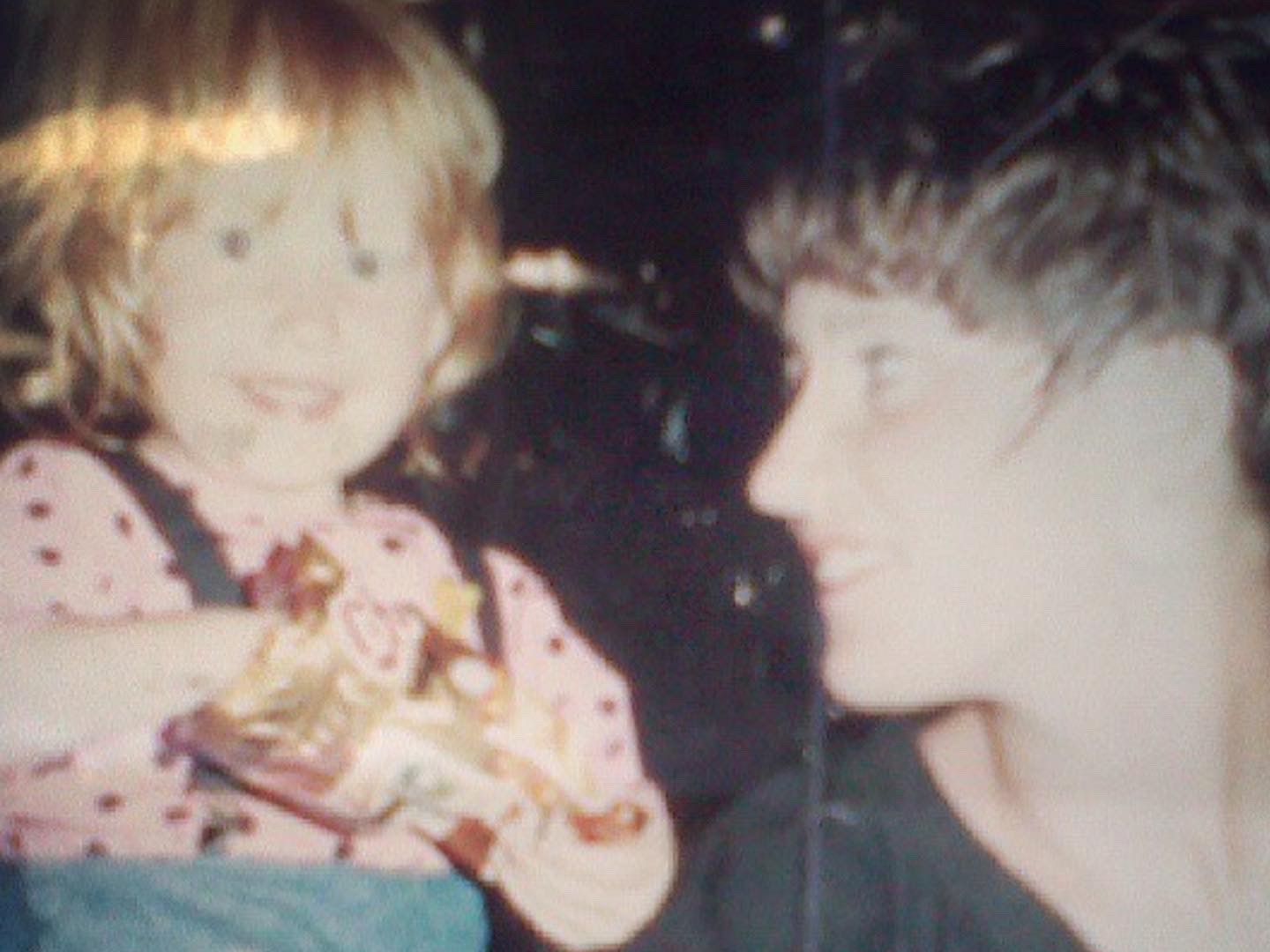 A toddler with ginger hair wearing a pink top eating a packet of crisps. Her mum, with brown hair and a black top, smiles at her