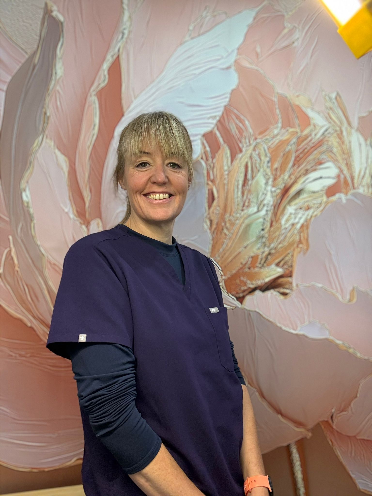 Portrait photo of a young woman wearing nurse scrubs in front of a painting of a pink flower.