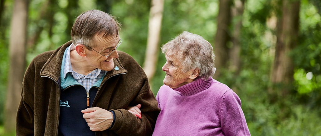 An elderly man and woman stroll together, sharing warm smiles as they walk side by side.