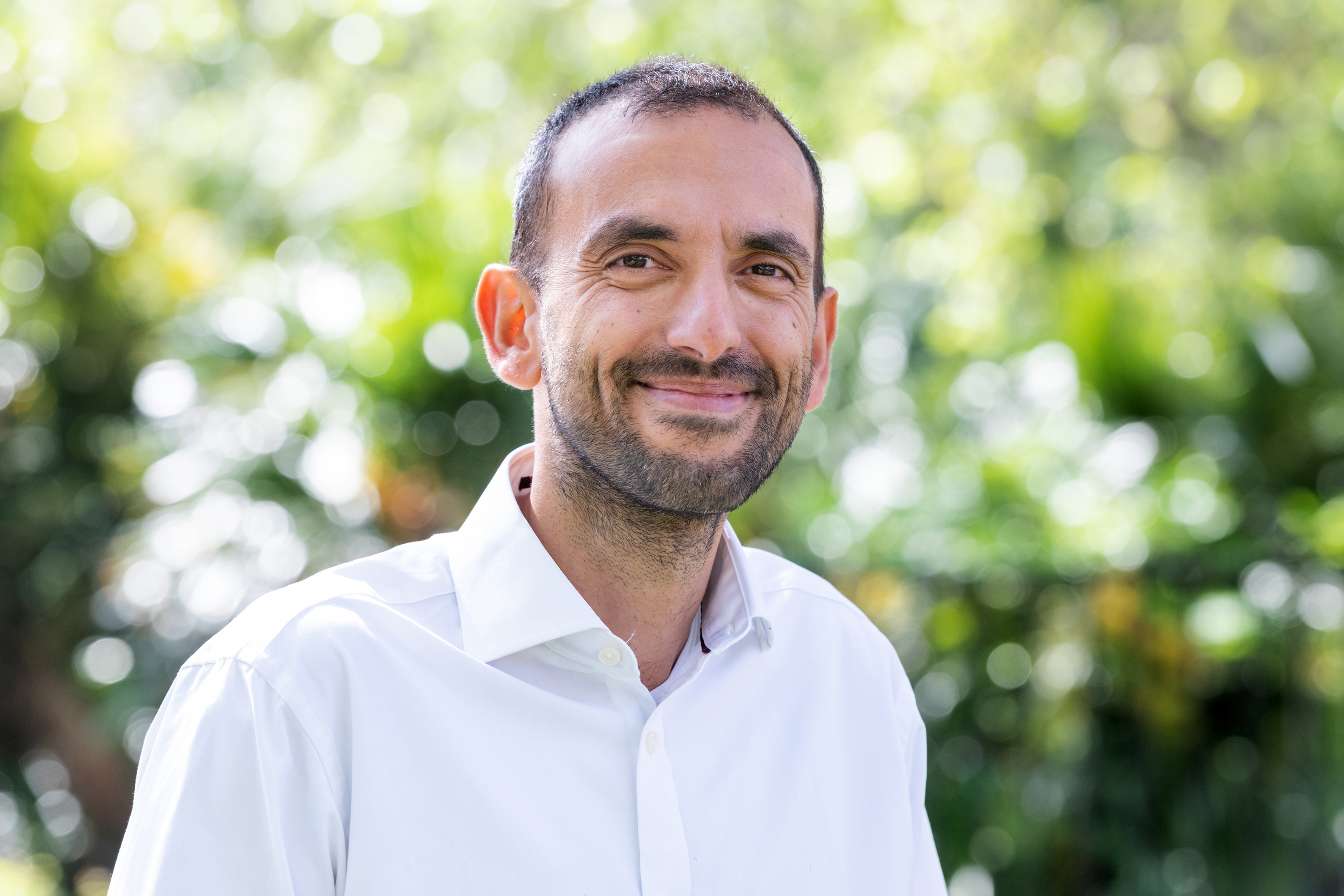 A man with short black hair and beard wearing a white shirt and smiling