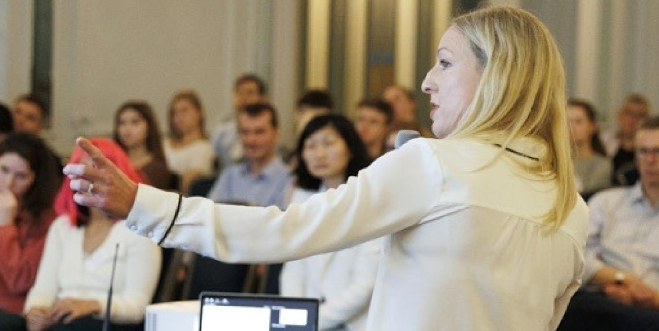 A woman stands before an audience delivering a presentation