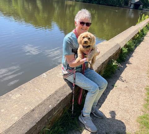 An elderly person with dementia sits by a river, holding her dog.
