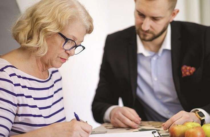 A man in a suit sits at a table with a woman, both are completing paperwork.