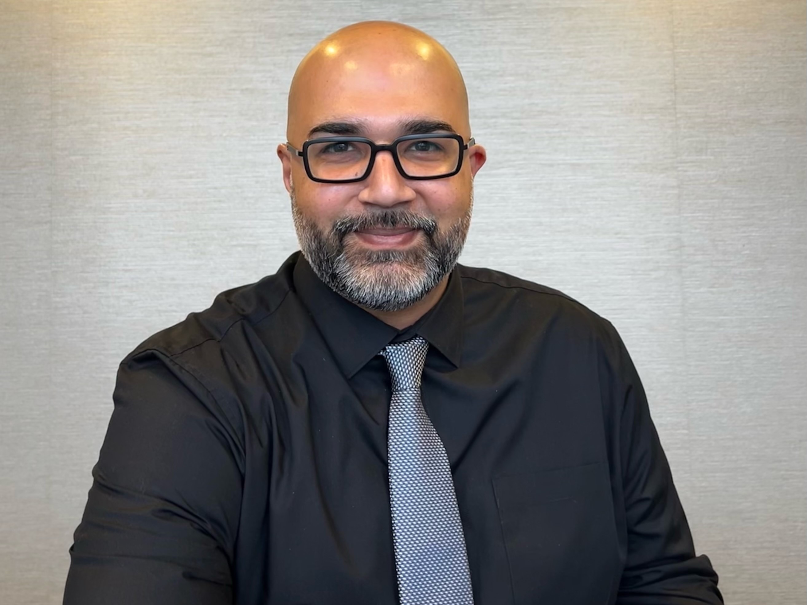 A smiling bald man with glasses and a beard, wearing a black shirt and grey tie