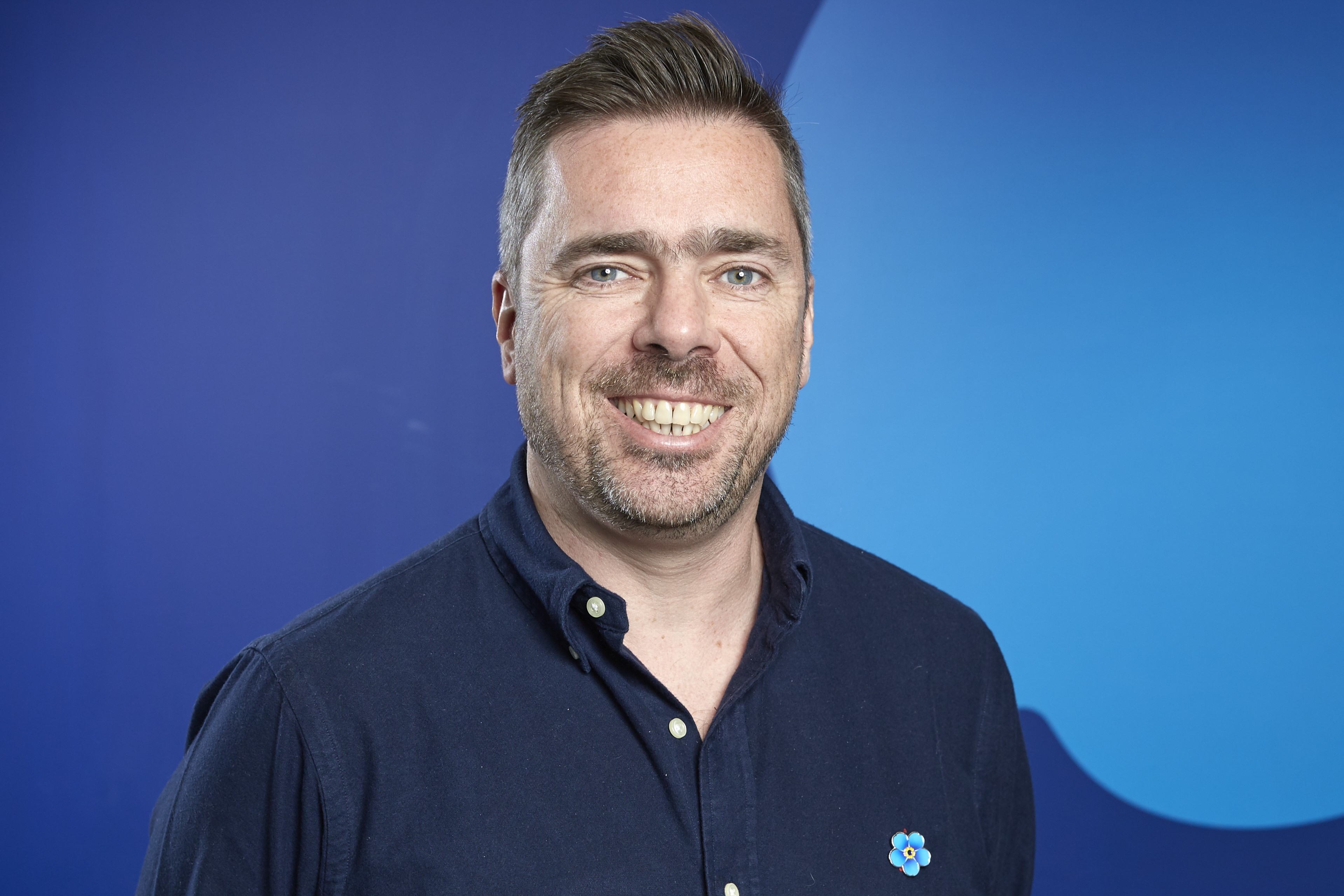 A man with short dark hair and a beard, wearing a blue shirt and smiling