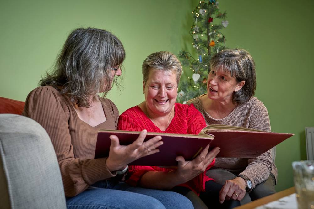 Mum and her two daughters smile as they flip through a photo album together, with a Christmas tree glowing in the background