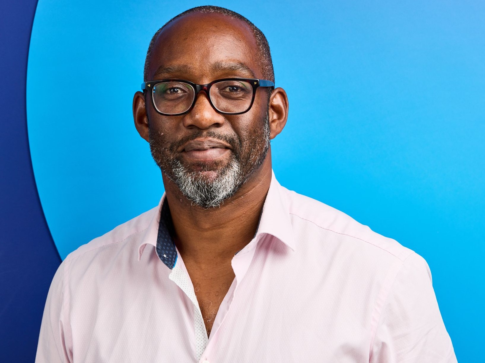A bald man with a black and white beard, black glasses and a light pink shirt