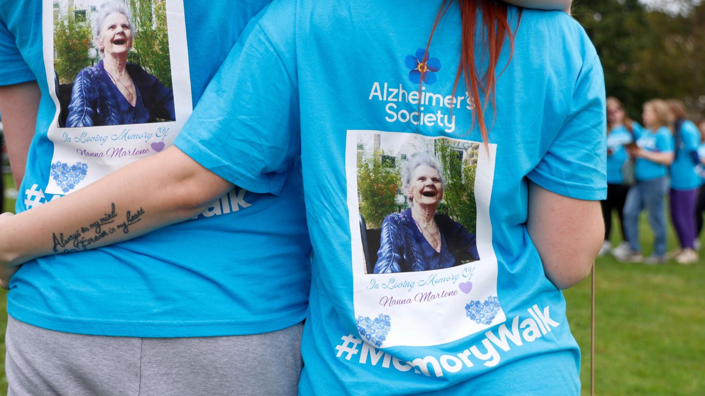 2 people arm in arm with their backs to the camera, wearing blue Memory Walk t-shirts with images of a loved one on the back.