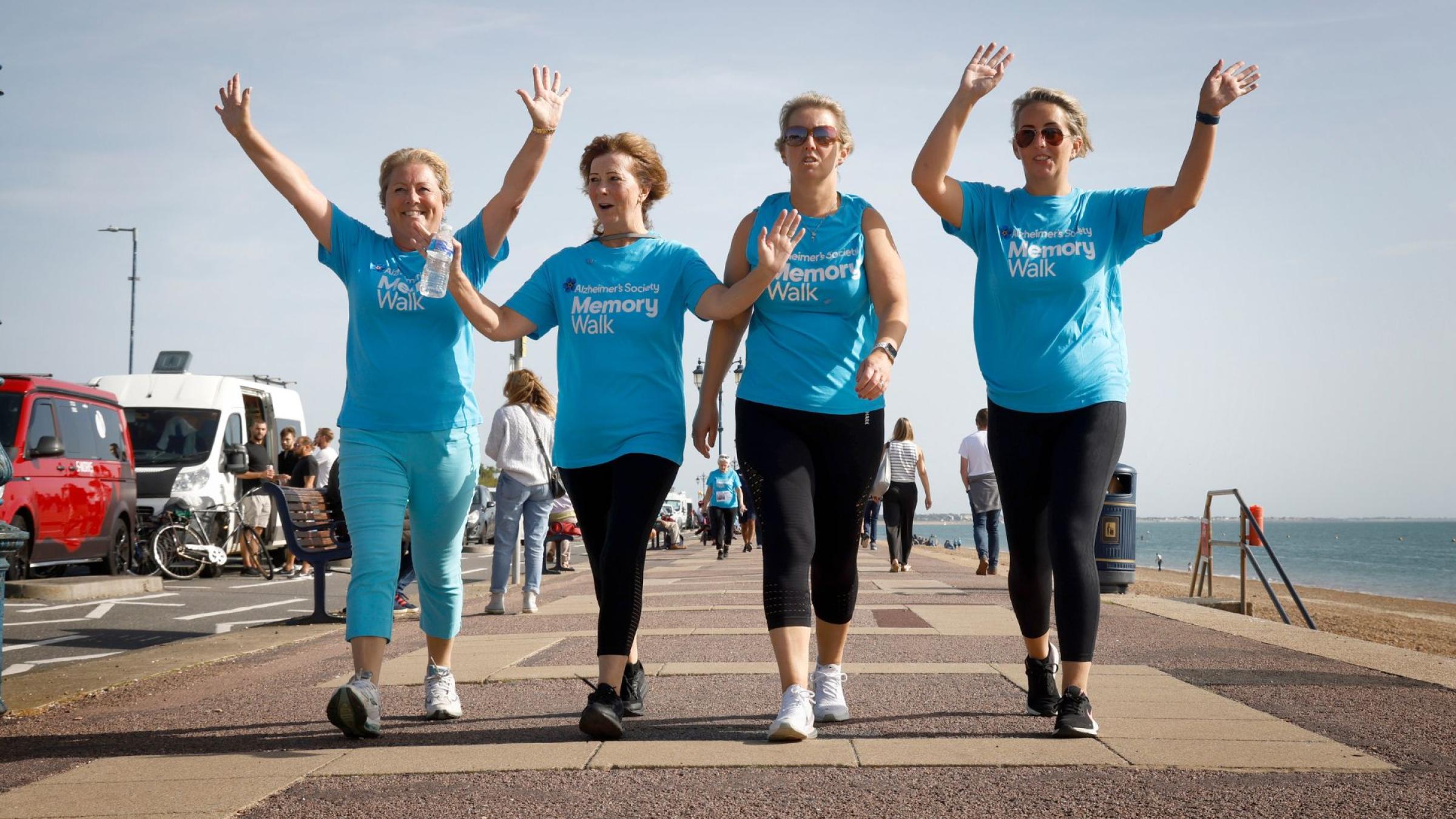 4 people in blue Memory Walk t-shirt, walking along the seafront with their arms in the air