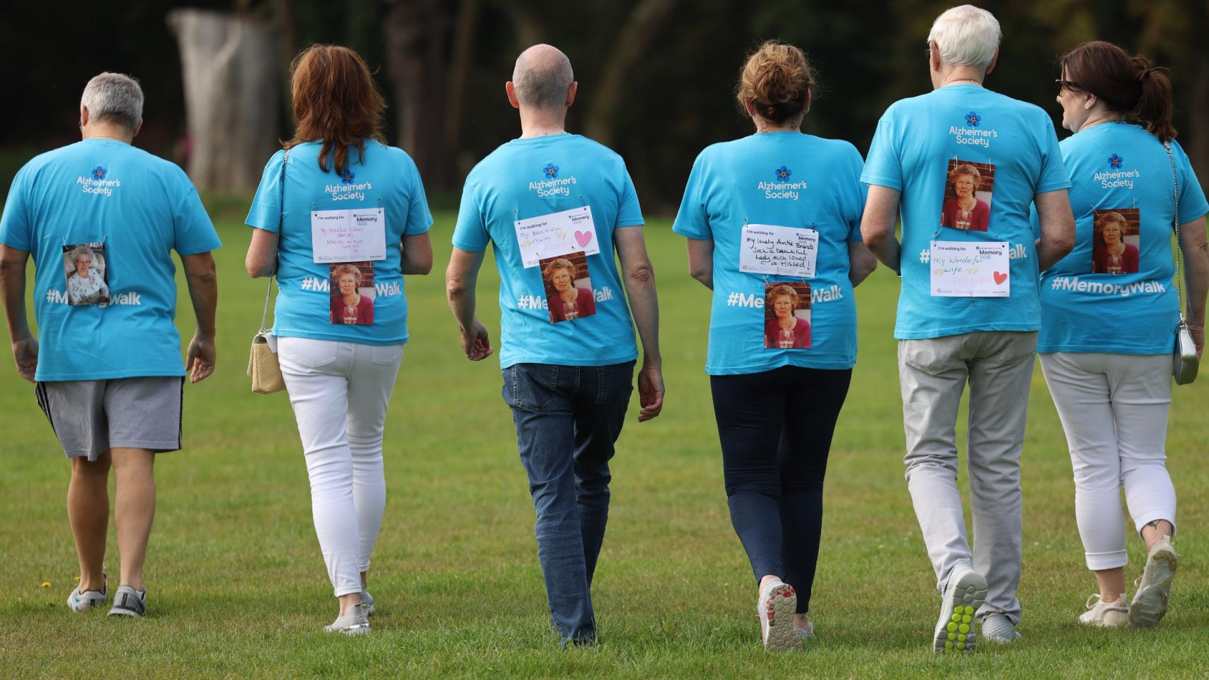 6 people in blue Memory Walk t-shirts, walking away from the camera with a picture of a loved one on their backs.