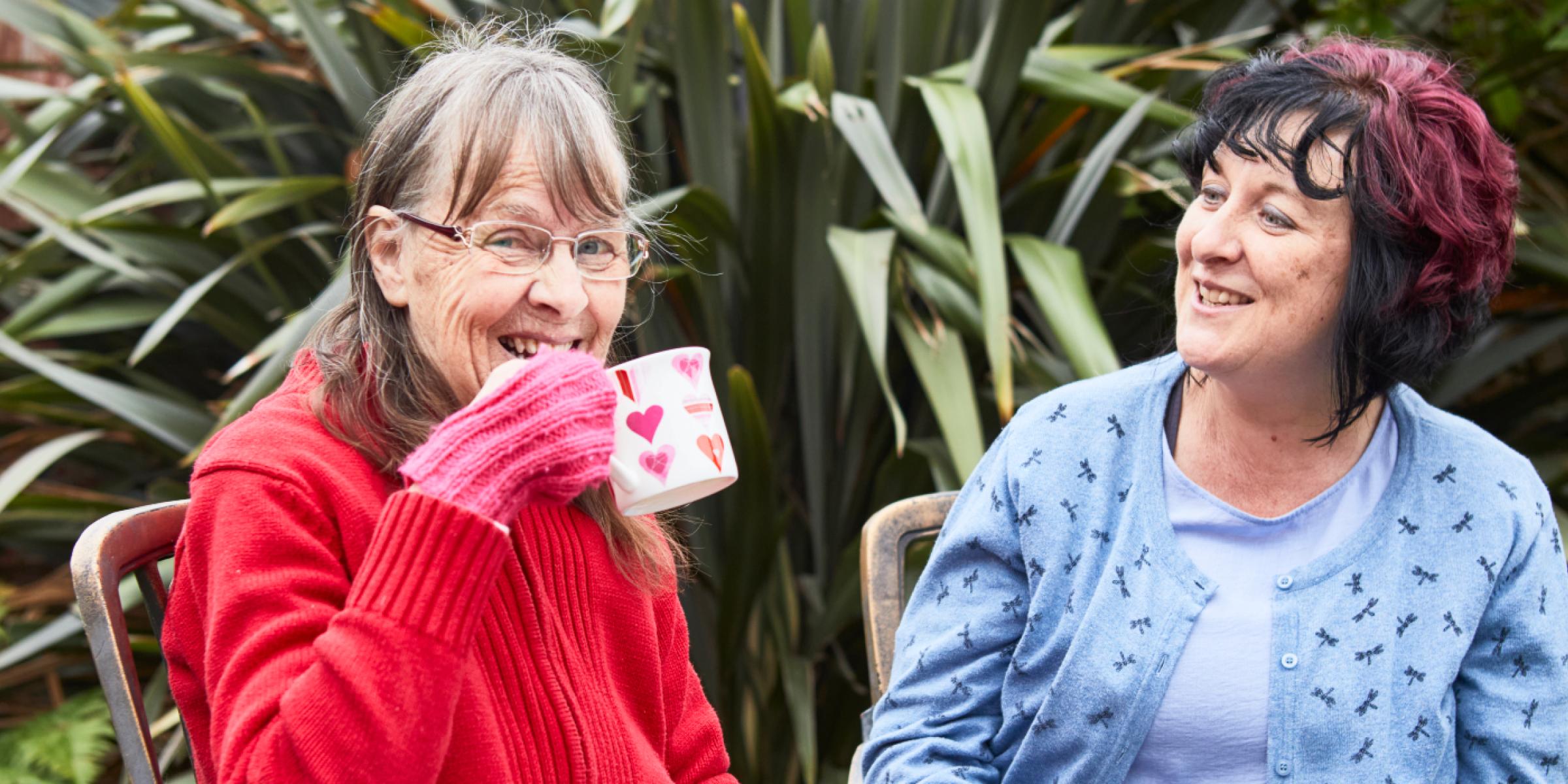 Louise and Marion sitting outside with a cup of tea wearing gloves and enjoying a conversation