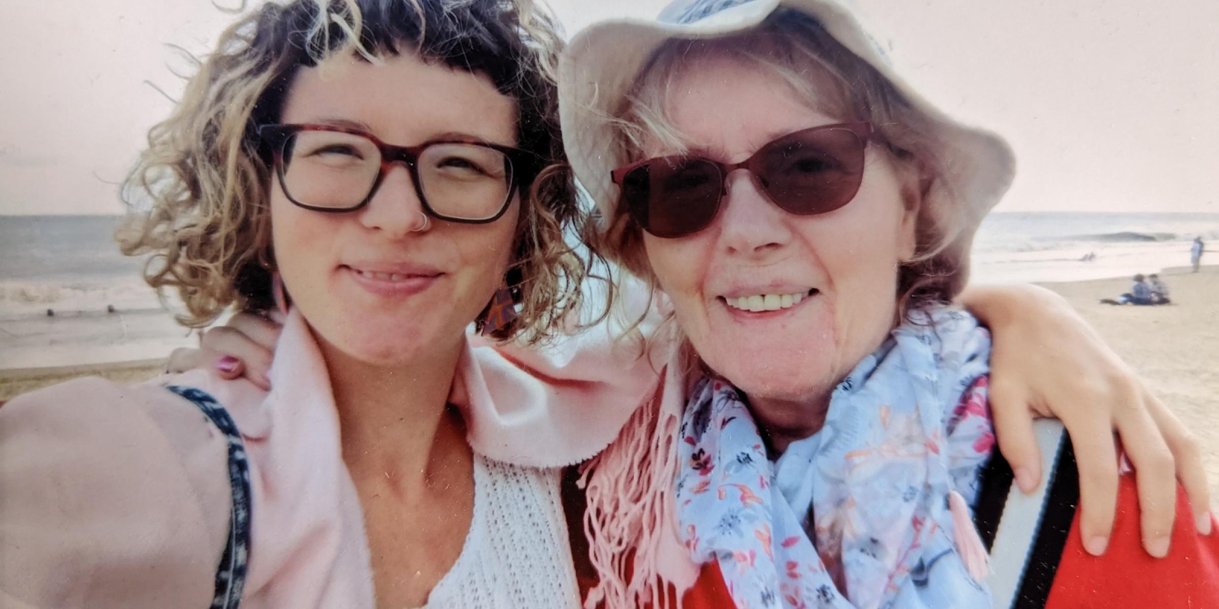 Cara has her arm around her mum while standing on a sandy beach and smiling together