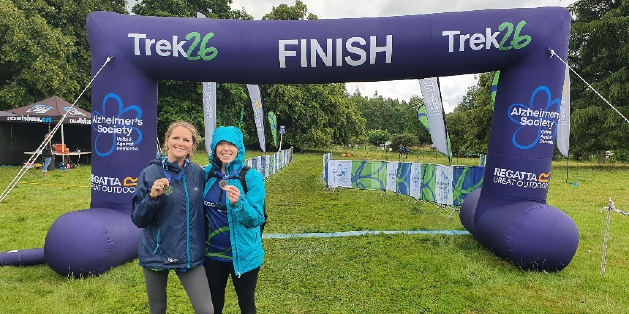 Julie and Karen under an inflatable arch that says 'Finish' at a Trek26 holding their medals