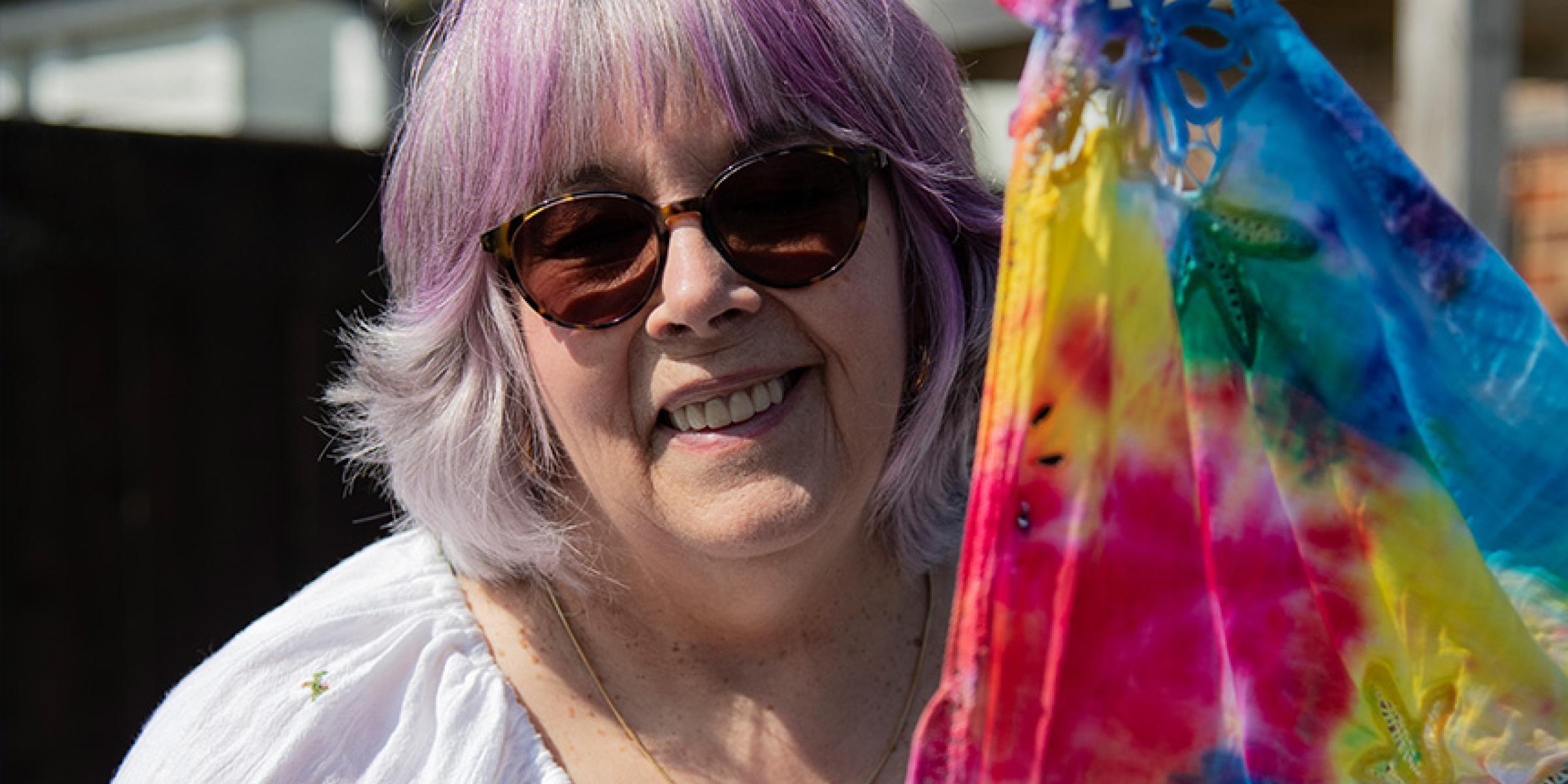 Gill Taylor stands outdoors next to some colourful tie-dyed fabric. She is wearing sunglasses and smiling.