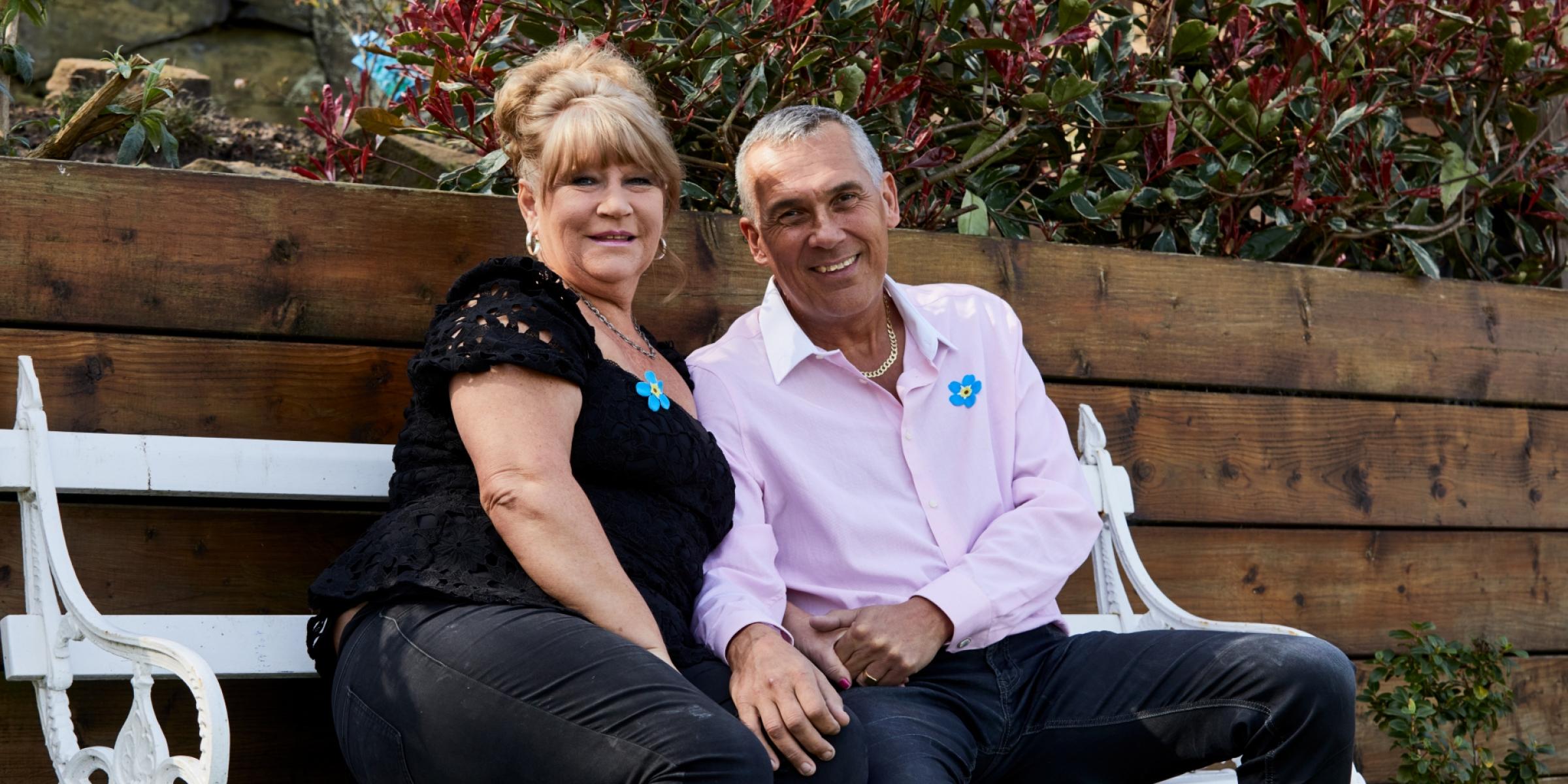 A woman and man sit on a garden bench smiling, wearing Alzheimer's Society Forget Me Not badges