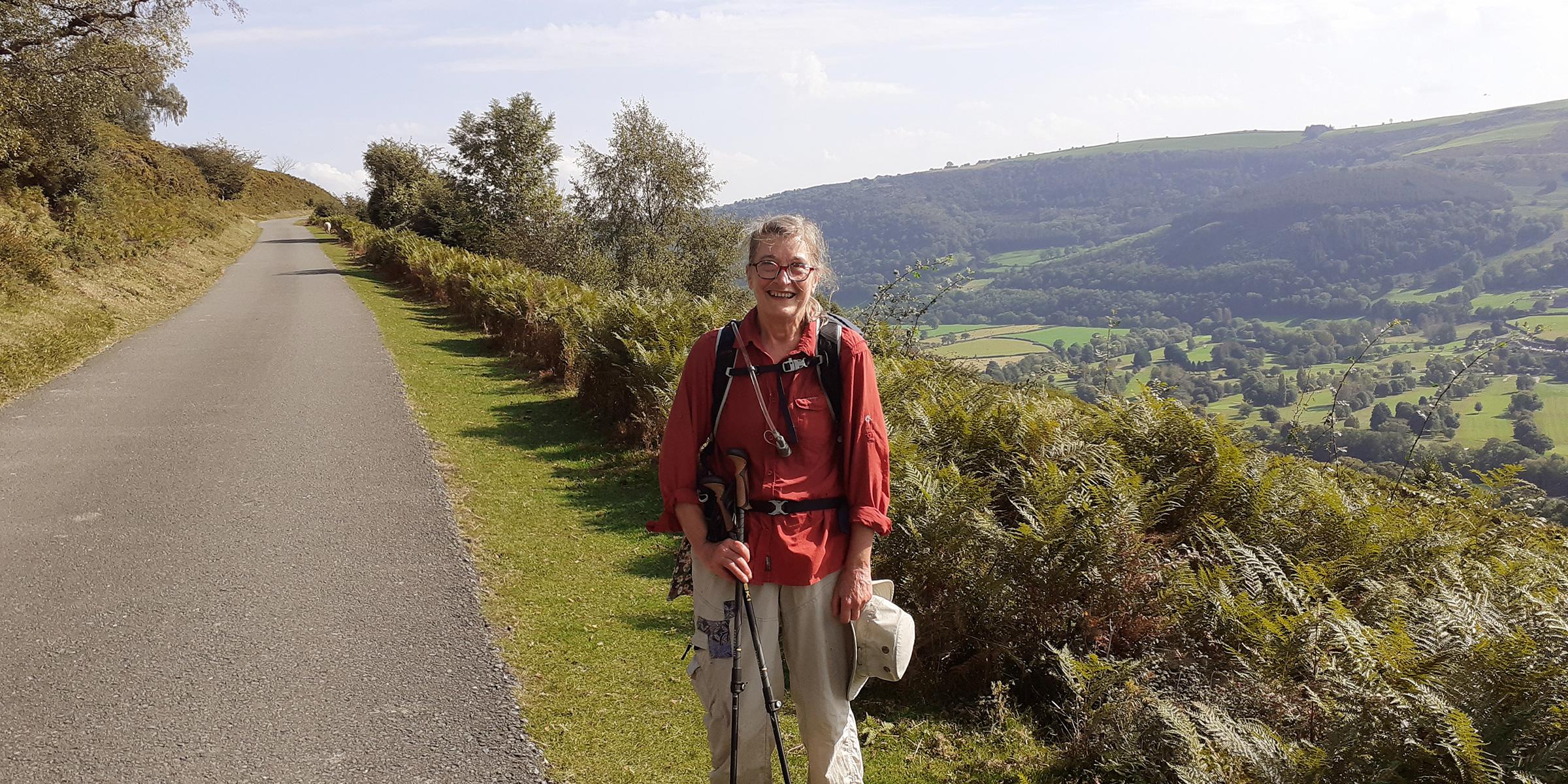 Jane Buckels stands along a road in front of a hilly area during her Offa's Dyke Path walk. She has a hat and walking stick and is wearing beige trousers and a red jumper