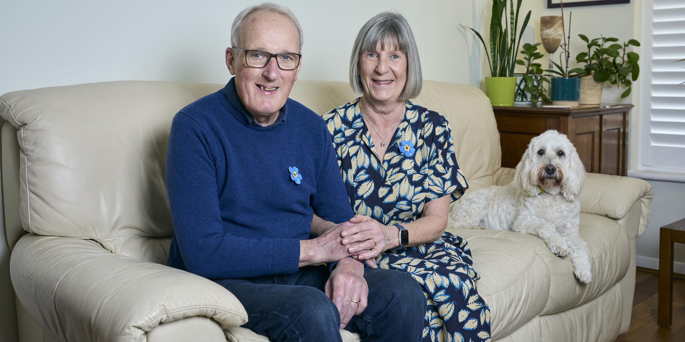 A man in a blue jumper, jeans and wearing glasses, a woman with a grey bob and a blue flowery dress and a white dog sit on a cream sofa