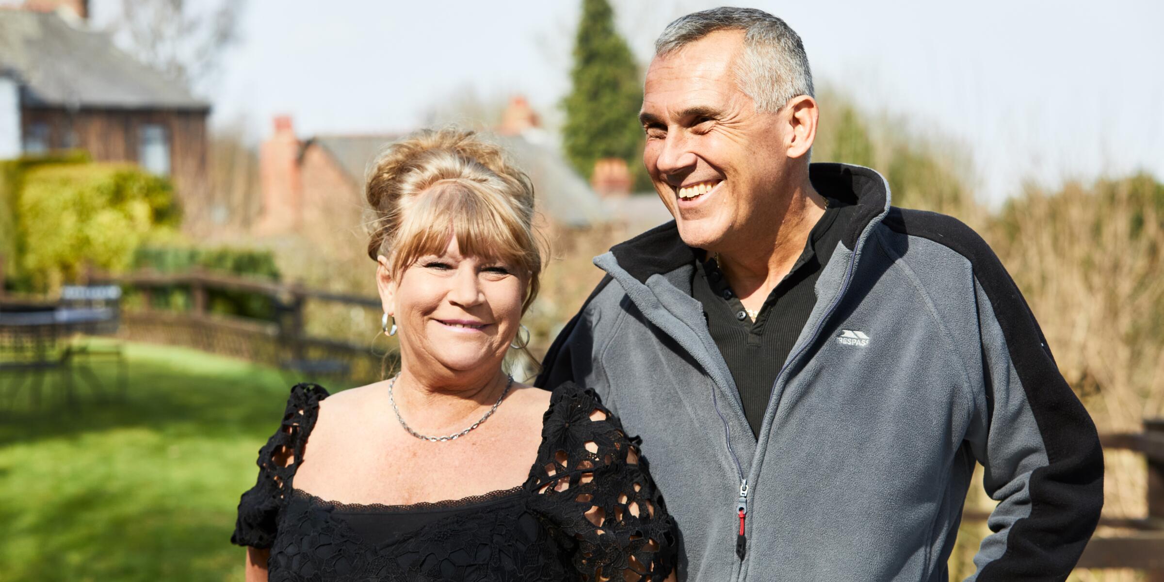 A woman with a blonde bun and black top and man with grey hair and a grey and black fleece smiling in a garden