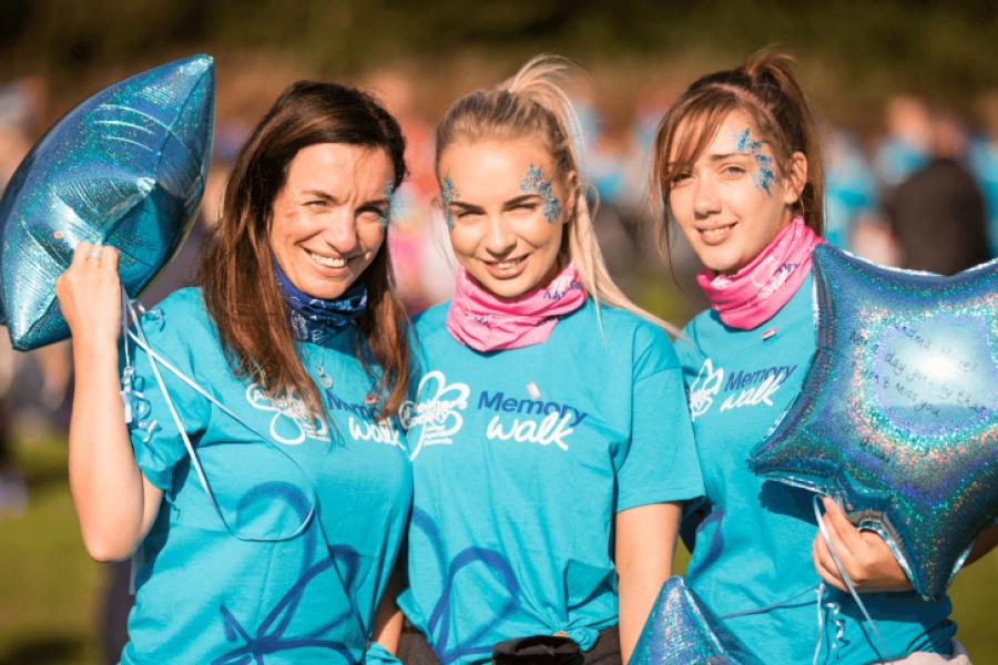 Three people wearing Memory Walk T-shirts and glittery face paint smile while holding blue star-shaped balloons