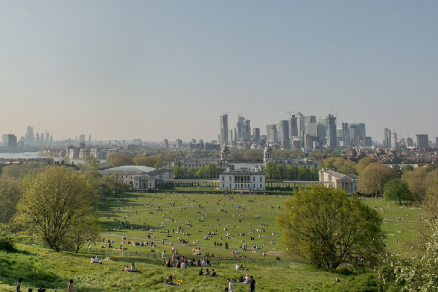A view of a London field in strong sunshine, people all across it sitting or sunbathing with London's tall buildings in the distance