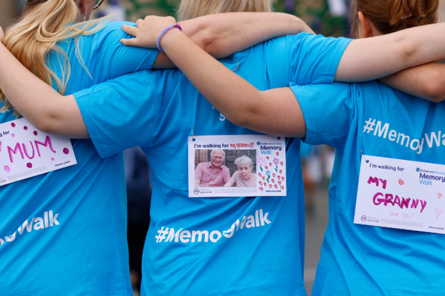 3 people arm in arm, with their backs to the camera. Each have a label on the backs of their blue Memory Walk t-shirts, saying they are walking for their mum/granny.