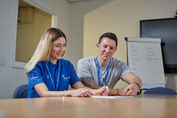 Two Telephone Dementia Advisers, one wearing a branded t-shirt sat at a desk smiling