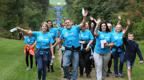A large group of people walking wearing Alzheimer's Society branded Memory Walk t-shirts walk towards to viewer, a procession of people also part of this Memory Walk can be seen from the foreground to far into the background