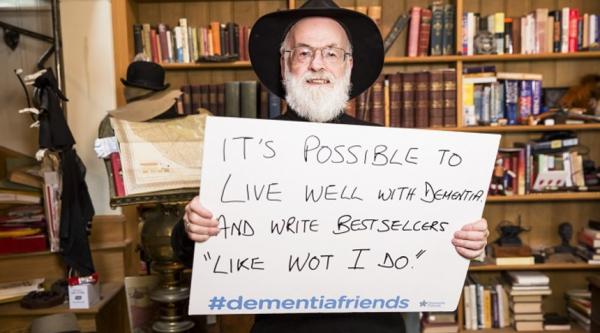 Terry Pratchett holding a sign which reads: 'It's possible to live well with dementia and write bestsellers "like wot I do"'