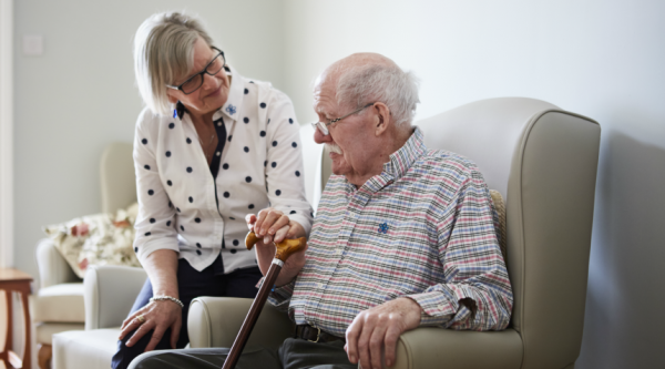 Woman looking lovingly at man sitting beside her in an armchair, holding a walking stick