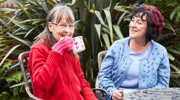 Louise and Marion with a cup of tea outside in the garden wearing gloves and smiling during a conversation