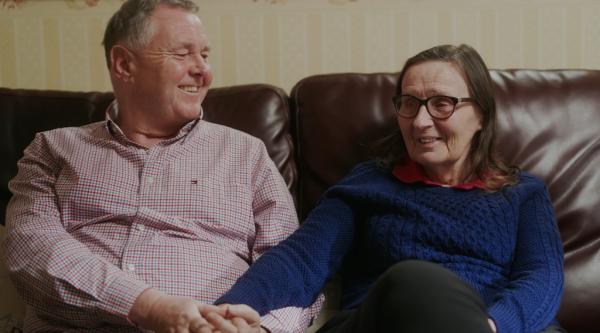 Bill and Jo sat on their couch, smiling and holding hands