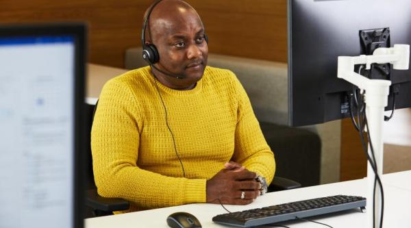 A Black person in a yellow jumper wearing a headset is listening to a phone call