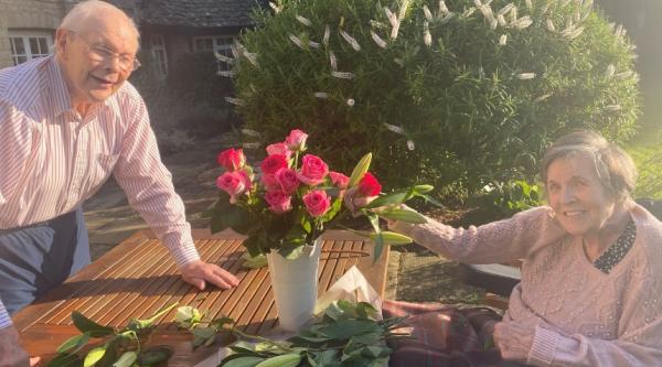 Carrie's mum and dad sit at a table in the garden, with a vase of flowers in the middle 