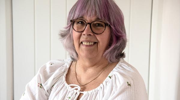 Gill Taylor is wearing glasses and a white blouse ad has purple hair. She stands in her home, cross armed, holding a paintbrush in her right hand, looking at the camera
