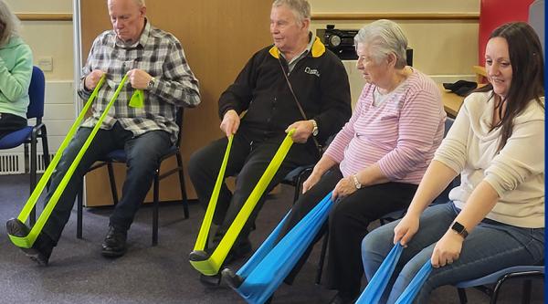 Four people participate in a Sit to Keep Fit session, they sit on chairs holding resistance bands which they stretch out with one leg and foot